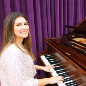 Smiling woman playing a wooden grand piano in front of purple stage curtains.
