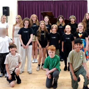 Group of children and teens posing in a music rehearsal hall; piano, purple curtain, and wooden floor visible.
