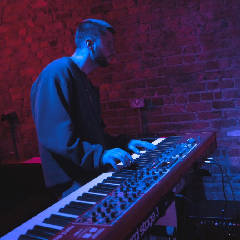 Man playing a red Nord Stage keyboard in a club with blue and magenta lighting against a brick wall.